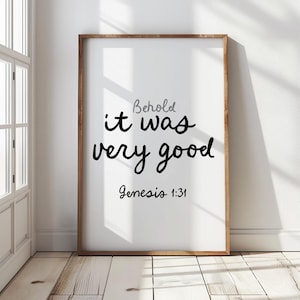 May include: A framed print with the words "Behold it was very good" in black script, with "Genesis 1:31" below. The frame is a warm brown, and the background is white. The print is displayed against a white wall and wooden floor.