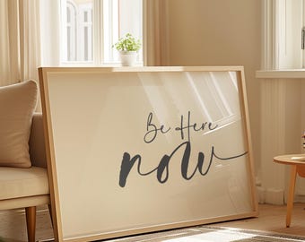 May include: A framed art print with the words "Be Here Now" in a stylish script. The artwork features a neutral colour palette with a light beige background and a wooden frame. The print is displayed in a living room setting.