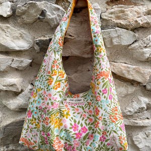 Può includere: Una borsa a spalla stile hobo con base in tessuto bianco e un motivo floreale colorato nei toni del rosa, arancione, giallo e azzurro, accentuato da fogliame verde. La fodera interna è arancione tinta unita e una piccola etichetta riporta Anne sy pique.
