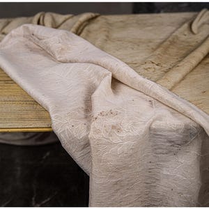 May include: A draped, off-white fabric with an embroidered floral pattern. The fabric is folded over a wooden surface, with a second, similar fabric in the background. The overall color palette is neutral, with subtle variations in tone.