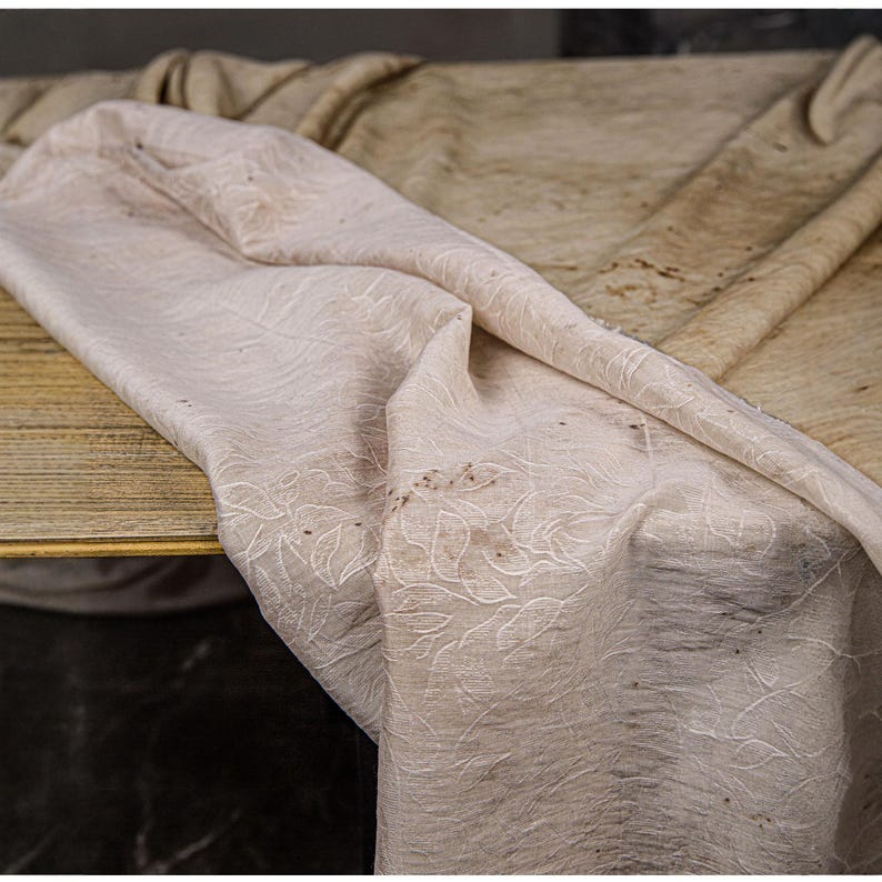 May include: A draped, off-white fabric with an embroidered floral pattern. The fabric is folded over a wooden surface, with a second, similar fabric in the background. The overall color palette is neutral, with subtle variations in tone.