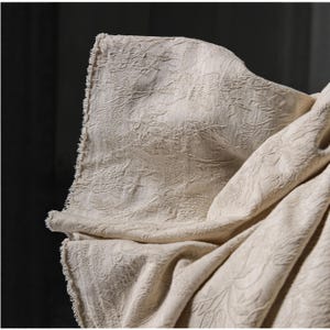 May include: Close-up of a cream-colored fabric with a detailed floral pattern. The fabric is draped, showing the texture and design. The edges have a fringed detail, adding to the visual interest. The background is dark, highlighting the fabric's texture.