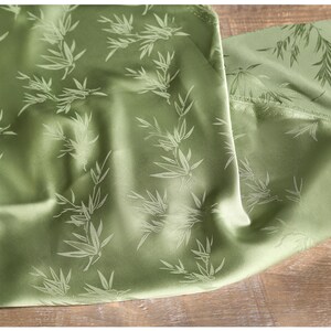 May include: Close-up of a green fabric with a bamboo leaf pattern. The fabric has a satin sheen and is folded, revealing the texture and design. The bamboo leaves are a lighter shade of green.