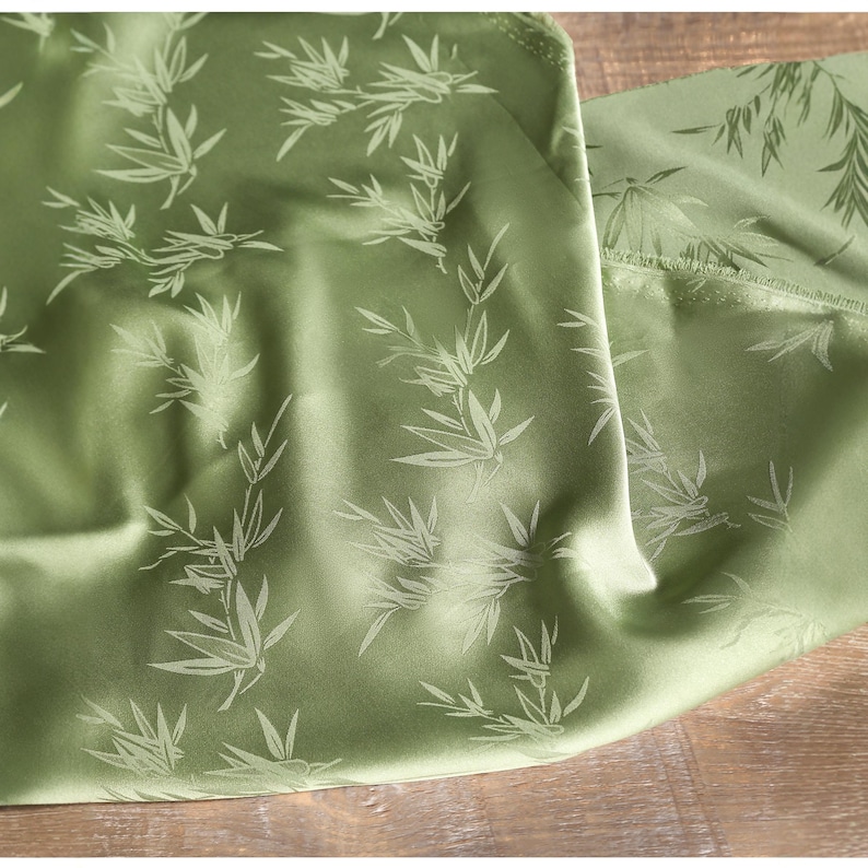 May include: Close-up of a green fabric with a bamboo leaf pattern. The fabric has a satin sheen and is folded, revealing the texture and design. The bamboo leaves are a lighter shade of green.