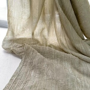 May include: A draped, semi-transparent fabric in olive green and beige tones. The material features a textured, vertical striped design, with folds and shadows. The fabric appears lightweight and airy, suitable for curtains or scarves. The fabric is likely made of a natural fiber.