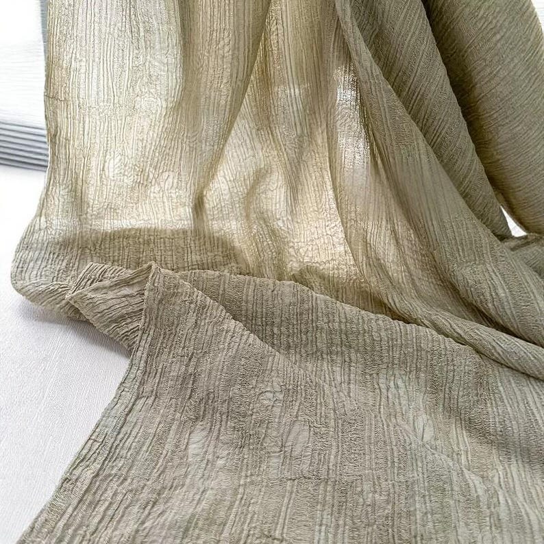 May include: A draped, semi-transparent fabric in olive green and beige tones. The material features a textured, vertical striped design, with folds and shadows. The fabric appears lightweight and airy, suitable for curtains or scarves. The fabric is likely made of a natural fiber.