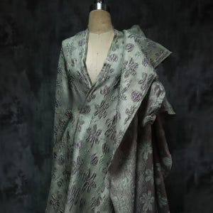 Puede incluir: Una prenda estilo envolvente de color verde salvia claro con un patrón floral texturizado en relieve en tonos morados y grises. La tela tiene una superficie arrugada y dimensional con escote en V y un panel drapeado asimétrico hacia un lado.