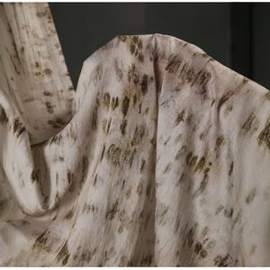 Puede incluir: Primer plano de una tela fluida con una est&eacute;tica natural y org&aacute;nica. El material es principalmente blanquecino con un patr&oacute;n de pinceladas y motas marrones y verde oliva. La tela se drapea suavemente, creando pliegues y sombras.