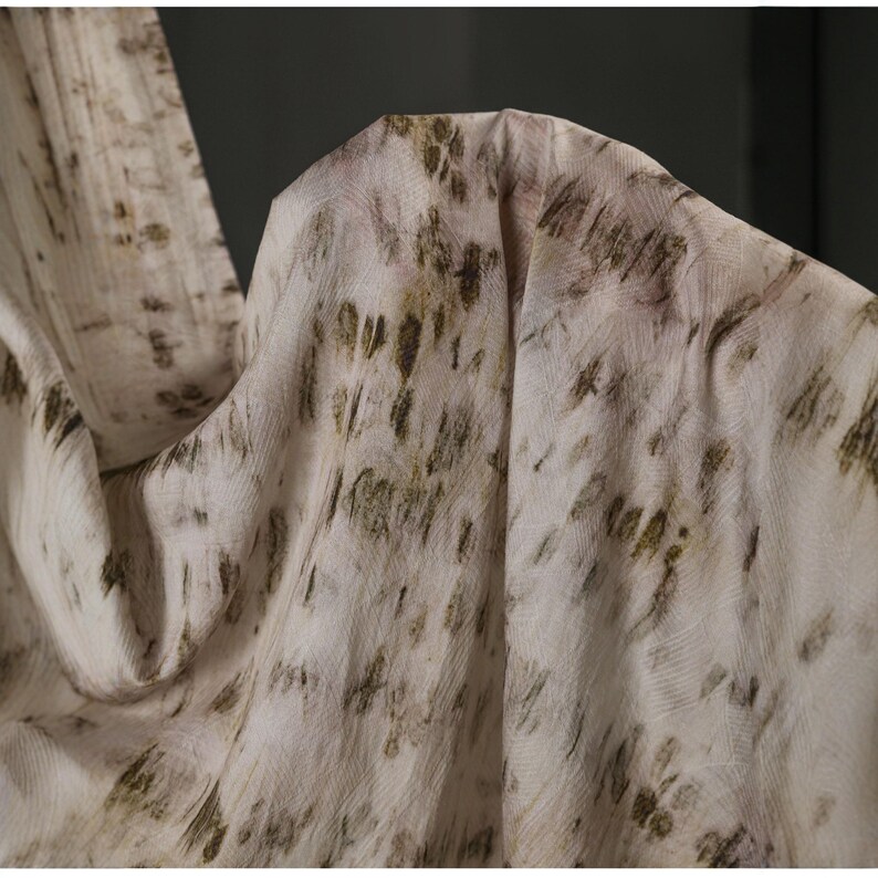 Puede incluir: Primer plano de una tela fluida con una est&eacute;tica natural y org&aacute;nica. El material es principalmente blanquecino con un patr&oacute;n de pinceladas y motas marrones y verde oliva. La tela se drapea suavemente, creando pliegues y sombras.