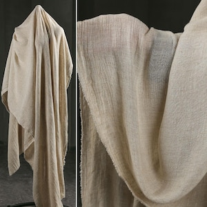 Può includere: Un tessuto beige drappeggiato, con un aspetto intrecciato e strutturato. Il tessuto è appeso, con pieghe e ombre che creano profondità. Il materiale appare morbido e leggero, adatto per abbigliamento o arredamento.