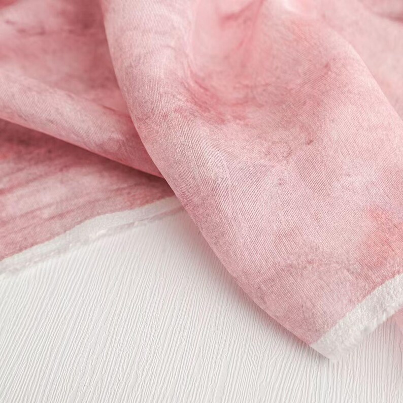 May include: A close-up of a pink fabric with a watercolor effect. The fabric is draped over a white textured surface. The pink color has variations, creating a soft, artistic look. The fabric's edge is finished with a white border.