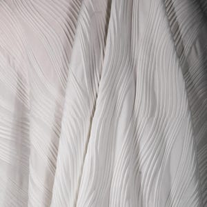 May include: Close-up of white fabric with a textured, wave-like pattern. The fabric appears to be folded, creating shadows and highlighting the texture. The material is a solid, neutral color, suitable for various applications.
