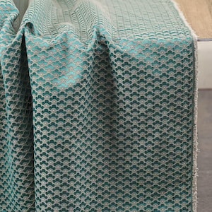 Op de afbeelding: Gevouwen teal stof met een herhalend geschulpt patroon. De stof heeft een getextureerde uitstraling en een gradiënteffect, van een lichter teal aan de bovenkant naar een donkerder teal aan de onderkant. De randen van de stof zijn gerafeld.