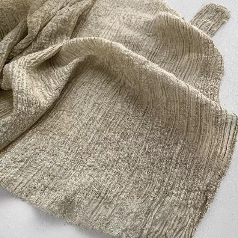 May include: Close-up of a beige fabric with a textured, crinkled appearance. The fabric is folded over itself, creating shadows and highlighting the detailed weave. The material appears soft and lightweight, suitable for clothing or home decor.