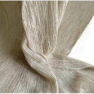 Puede incluir: Primer plano de una tela fluida y texturizada en color beige claro. El material tiene un aspecto arrugado con l&iacute;neas verticales y paralelas, creando un efecto suave y drapeado. Los pliegues y curvas de la tela son visibles, lo que sugiere un textil ligero.
