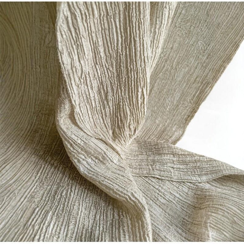 Puede incluir: Primer plano de una tela fluida y texturizada en color beige claro. El material tiene un aspecto arrugado con l&iacute;neas verticales y paralelas, creando un efecto suave y drapeado. Los pliegues y curvas de la tela son visibles, lo que sugiere un textil ligero.