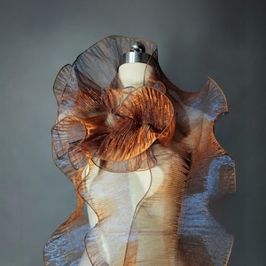Puede incluir: Una prenda con volantes de color cobre se exhibe en un maniquí. La tela tiene un brillo metálico y está en capas para crear un efecto voluminoso. El diseño presenta un gran detalle en forma de flor en el centro y una forma asimétrica.