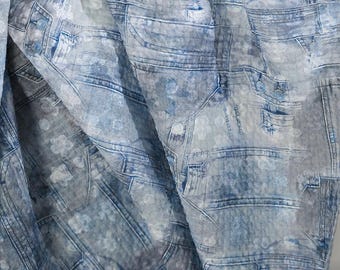 May include: A fabric with a denim print in shades of blue and gray. The design mimics the look of jeans, with details like pockets, seams, and button closures. The fabric is folded, creating shadows and depth.