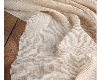 Può includere: Tessuto color crema, strutturato con dettagli a coste verticali. Il tessuto è drappeggiato e piegato, creando ombre e luci soffuse. Il materiale sembra naturale, forse lino o cotone, ed è posto su una superficie di legno.