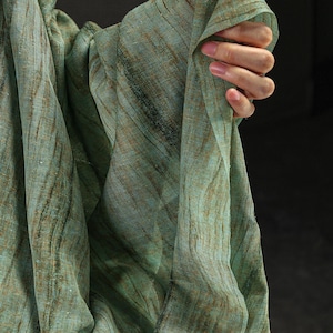 以下が含まれることがあります： 緑と茶色の色合いの、流れるようなシアー生地。素材は、微妙な濃い線が走るテクスチャード加工が施されています。生地はドレープされており、柔らかい折り目と動きの感覚を作り出しています。全体的な美しさは自然で有機的です。