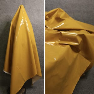 May include: Mustard yellow, shiny fabric draped over a surface. The fabric has a glossy, reflective finish, with folds and creases creating shadows. The material appears to be a smooth, synthetic textile, possibly vinyl or a similar coated material.