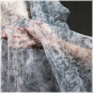 Puede incluir: Una tela transparente con un patrón tipo acuarela en tonos grises y blancos. La tela está drapeada, con una mano visible a través del material translúcido. El diseño se asemeja a un cielo nublado o una textura de mármol.