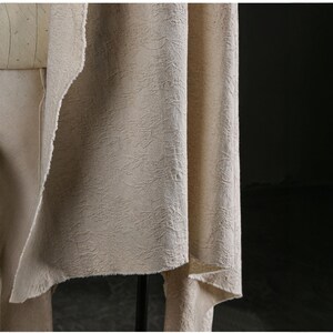 May include: Close-up of beige fabric with a textured, organic pattern. The fabric is draped, revealing a raw, unfinished edge. The neutral color and subtle texture suggest a natural fiber textile, possibly for clothing or home decor.