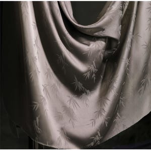 May include: A draped, silky fabric in a muted gray-green hue. The material features a subtle pattern of bamboo leaves. The fabric is smooth and reflects light, creating highlights and shadows that emphasize its texture and drape.