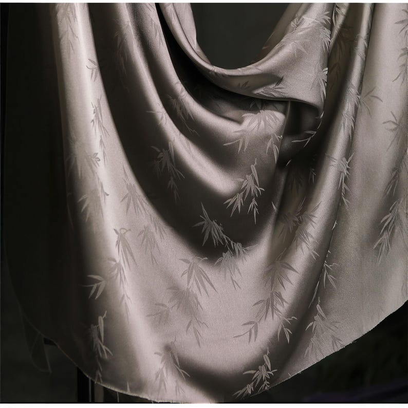 May include: A draped, silky fabric in a muted gray-green hue. The material features a subtle pattern of bamboo leaves. The fabric is smooth and reflects light, creating highlights and shadows that emphasize its texture and drape.