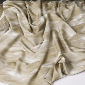 Może przedstawiać: Płynna tkanina z marmurkowym wzorem w odcieniach beżu i bieli. Materiał jest udrapowany, tworząc miękkie fałdy i cienie. Tkanina wydaje się lekka i nadaje się do odzieży lub dekoracji wnętrz.
