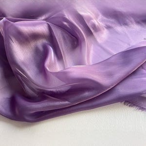 Puede incluir: Primer plano de una tela fluida y transparente en tonos lavanda y morado. La tela tiene un brillo brillante, con la luz reflejándose en los pliegues y creando reflejos. Los bordes de la tela están ligeramente deshilachados.