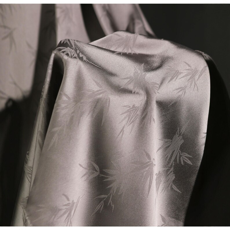 May include: A close-up of a gray fabric with a subtle sheen. The fabric is patterned with a repeating bamboo leaf design. The folds and draping of the fabric are visible, creating shadows and highlights.