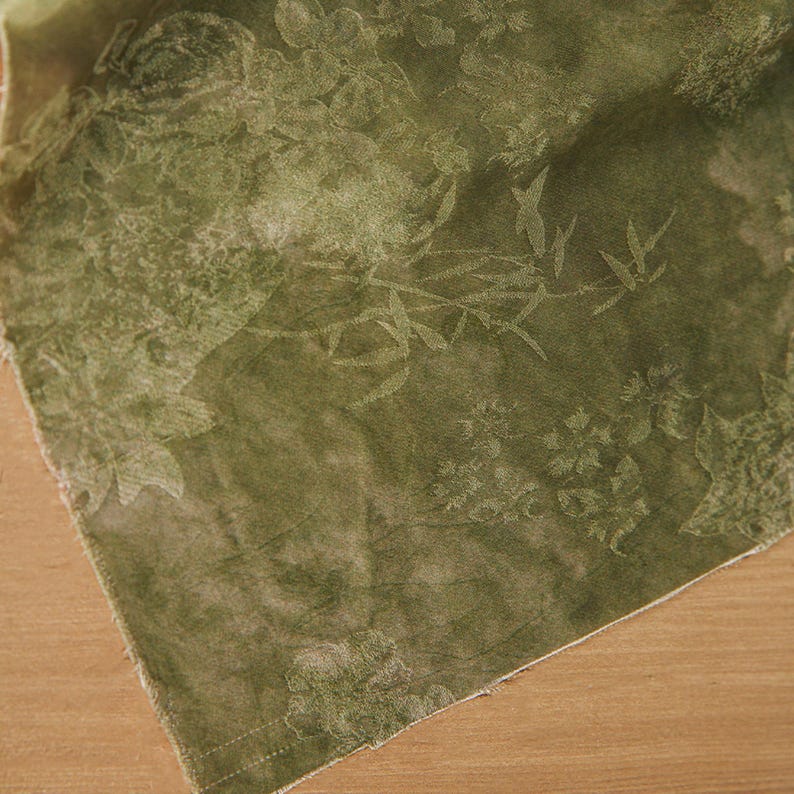 May include: Olive green fabric with a floral and bamboo leaf pattern. The fabric has a soft, textured appearance and is folded, revealing a glimpse of the raw edge. The design features detailed botanical elements.