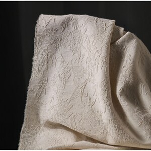 May include: Close-up of a beige fabric with a floral pattern. The fabric is folded, revealing its texture and design. The pattern features detailed leaf and vine motifs, creating a textured surface. The fabric's neutral color makes it suitable for various applications.