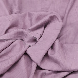 Pode incluir: Uma peça de tecido roxo claro com uma textura subtilmente enrugada e linhas horizontais visíveis em toda a superfície. O material está drapeado em dobras suaves, exibindo o acabamento mate e a cor lavanda consistente e suave.