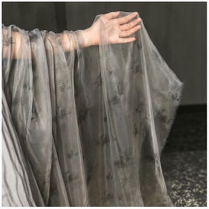 Peut inclure: Un tissu gris transparent avec un motif floral est maintenu, mettant en valeur sa texture délicate. Le tissu semble léger et fluide, avec un éclat subtil. Les bords sont inachevés, ajoutant à sa qualité éthérée. L'effet global est doux et élégant.