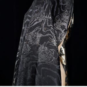 May include: A close-up of a black fabric with a floral pattern and silver accents. The fabric has a textured appearance and is draped, revealing a gold-colored lining. The background is black.