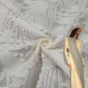 Puede incluir: Un vestido largo de color crema con mangas largas y un acabado texturizado de flecos deshilachados. La prenda incluye un sombrero de ala ancha en un tono crema a juego, creando un estilo monocromático con una tela suave y fibrosa.