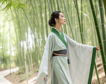 Könnte beinhalten: Eine Person trägt ein hellgrünes und weißes traditionelles Outfit im asiatischen Stil mit grünen Verzierungen und steht in einem Bambuswald. Das Outfit hat lange Ärmel und einen schwarzen Gürtel. Der Hintergrund zeigt einen Pfad und viele Bambusstämme.