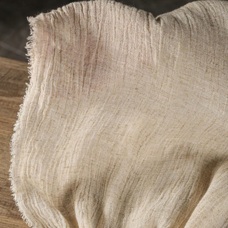 May include: Close-up of a beige linen fabric with a textured, wrinkled appearance. The fabric has a raw, unfinished edge on the left side. The material appears soft and lightweight, with subtle variations in color and tone.