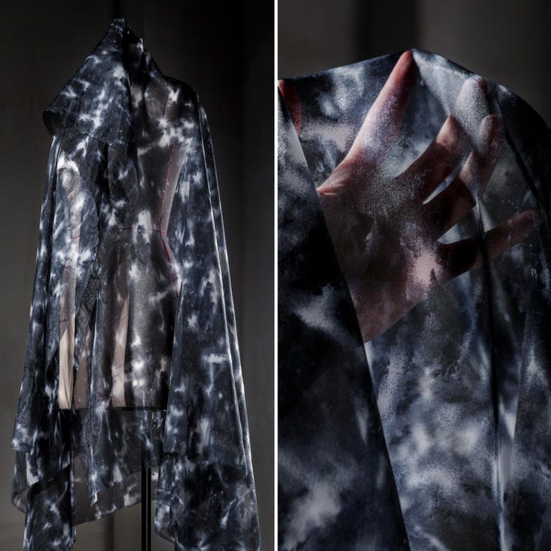 May include: A sheer, tie-dye fabric draped over a dress form. The fabric has a pattern of white and grey splotches against a dark background. The fabric is lightweight and appears to be flowing. The image is a close-up of the fabric.