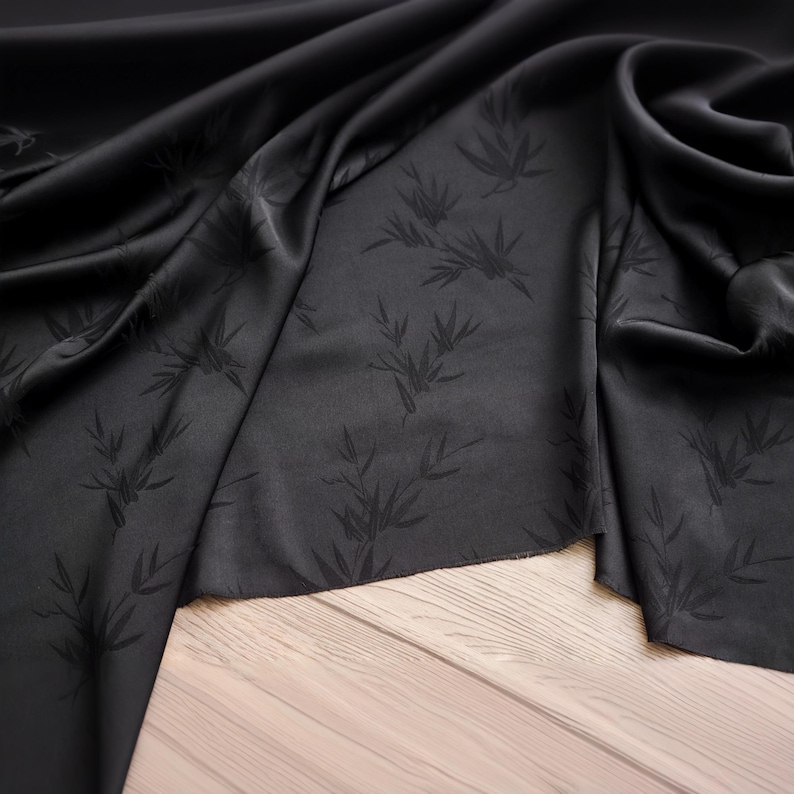 May include: Black fabric with a subtle sheen, featuring a repeating pattern of bamboo leaves. The fabric is draped, creating soft folds and shadows. The dark color and delicate pattern suggest a luxurious textile.