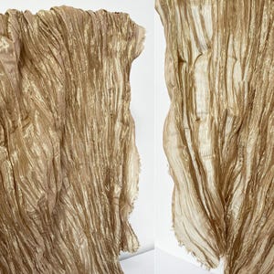 Puede incluir: Una bufanda de color dorado metálico que presenta una textura muy arrugada y plisada en toda la tela. El material tiene una calidad brillante y reflectante con líneas verticales y bordes deshilachados a los lados del tejido.