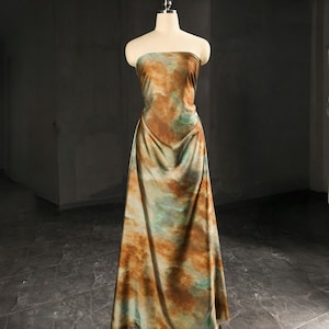 Puede incluir: Vestido largo sin tirantes sobre un maniquí. El vestido presenta un patrón tipo acuarela en tonos marrón, verde y azul. La tela se drapea elegantemente, mostrando la silueta fluida del vestido. Una prenda única.