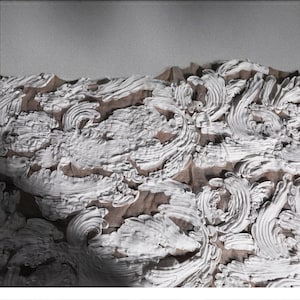 May include: Close-up of a textured artwork featuring white, swirling, raised patterns against a brown background. The design has a three-dimensional effect, with the white elements appearing to be layered and sculpted. The artwork is framed.