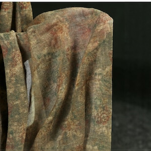 May include: Close-up of a draped fabric in muted tones. The textile displays a mix of olive green, brown, and reddish-brown shades, with delicate floral or patterned elements. The fabric seems lightweight and slightly transparent, with a textured finish.