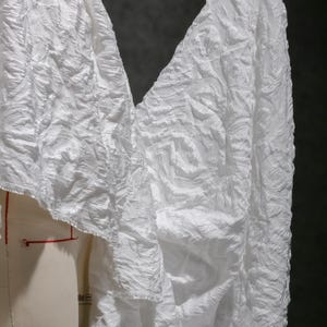 Może przedstawiać: Zbliżenie białej tkaniny o teksturowanym, wirującym wzorze. Tkanina jest udrapowana na manekinie, prezentując jej płynną jakość. Materiał wydaje się lekki i odpowiedni do odzieży.