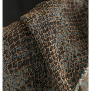 Puede incluir: Primer plano de una tela texturizada con un patrón de escamas de reptil. La tela presenta una combinación de colores marrón oscuro y verde azulado, con un diseño tejido. El material está doblado, revelando la textura y las variaciones de color.
