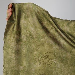 May include: Olive green fabric with a floral and bamboo leaf pattern. The fabric is held up, revealing its texture and design. The pattern is a lighter shade of green, creating a subtle contrast against the darker background.
