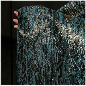 Puede incluir: Primer plano de una tela texturizada con un patrón en verde azulado, negro y dorado. La tela tiene un diseño irregular en relieve, creando un efecto brillante. El material se sostiene, mostrando su caída y textura.